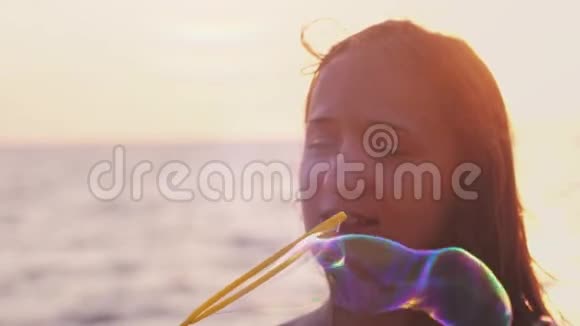 年轻快乐的黑发女人在美丽的夕阳下在海滩上吹着肥皂泡在海边缓慢地微笑着视频的预览图