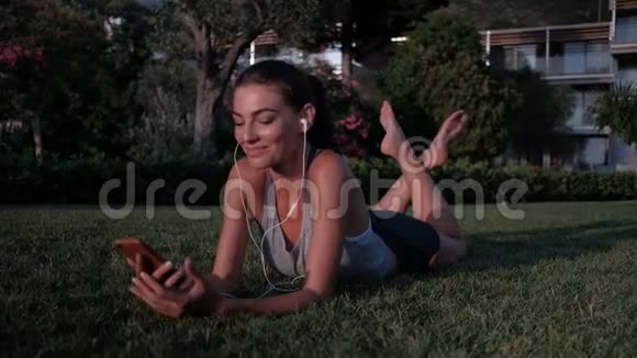 躺在草地上的女人用电话在耳机上听音乐视频的预览图