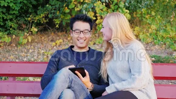 漂亮的年轻男女坐在公园的长凳上享受数码平板电脑吧韩国男人和高加索女人视频的预览图