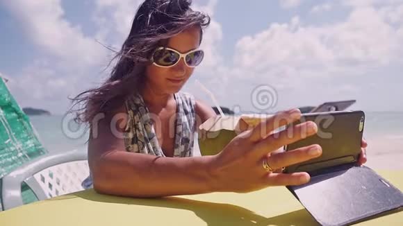 年轻的黑发女人戴着太阳镜通过电话自拍喝着椰子做的鸡尾酒心情轻松视频的预览图