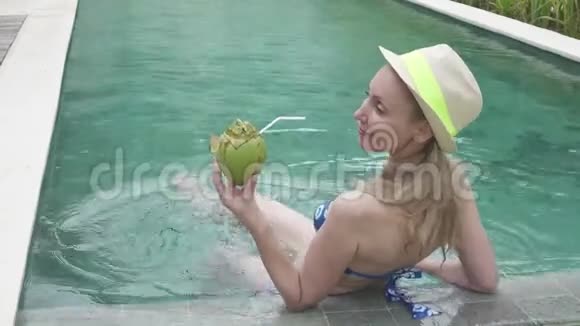 比基尼的年轻苗条的女人喝着热带度假胜地游泳池边缘的可可椰子汁视频的预览图