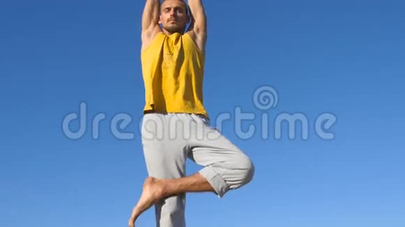 年轻运动男子站在树瑜伽姿势的肖像以蓝天为背景高加索人练习瑜伽动作视频的预览图