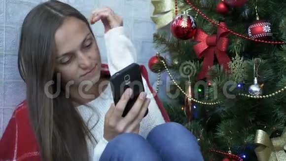 一个女人坐在一个有红色格子的地板上在圣诞树旁边使用智能手机视频的预览图