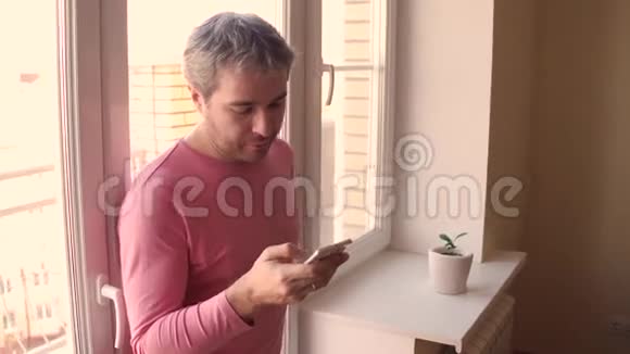 帅哥穿着粉红色T恤滚动应用程序在他的手机附近的大窗户4K近距离射击视频的预览图