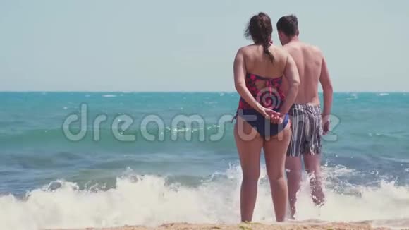 海滩夫妇从后面看海景一对夫妇站在白色的沙滩上度假海浪飞溅视频的预览图