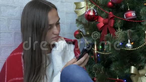 一个女人坐在一个有红色格子的地板上在圣诞树旁边使用智能手机视频的预览图