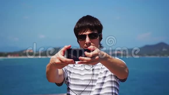 戴太阳镜的年轻人带着他的智能手机在海滩上自拍用耳机听音乐苏梅岛视频的预览图