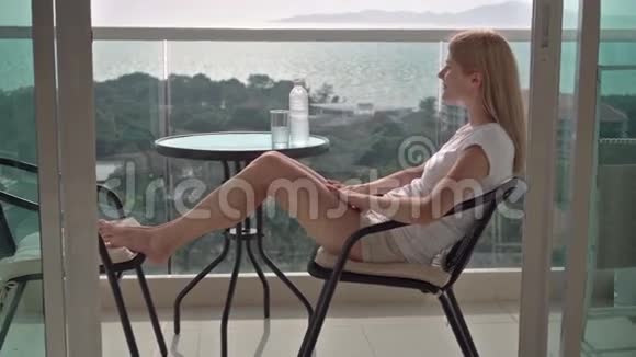 美丽迷人的女人坐在阳台的椅子上放松享受风景饮用水视频的预览图