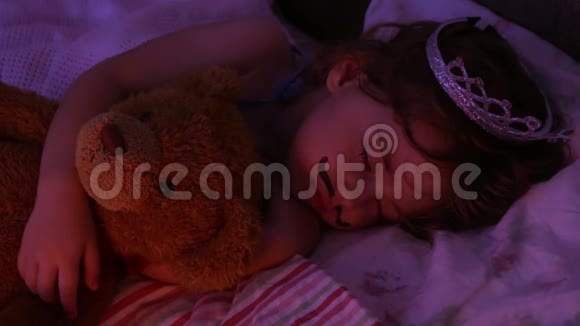 特写肖像小女孩睡在床上戴着公主王冠的女孩抱着一个玩具熊玩具在床上视频的预览图