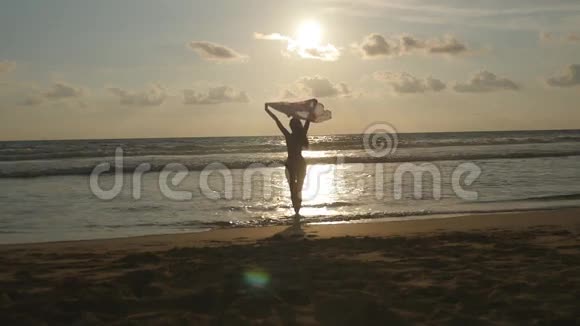 日落时分戴着围巾的年轻幸福的女人站在海边喜欢夏天的女孩比基尼女性剪影视频的预览图