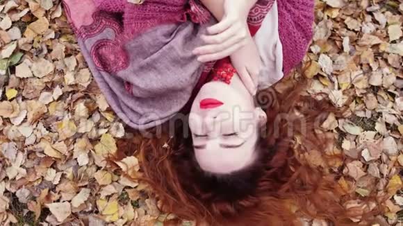 在秋天的森林里一头红色卷发的漂亮女孩在干燥的叶子上缓慢地躺着3840x21604k视频的预览图