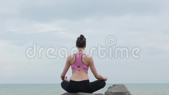 女孩练习瑜伽健身运动靠近海边坐在石头上的莲花姿势视频的预览图