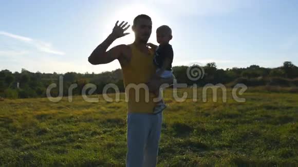 在阳光明媚的夏日年轻的父亲在大自然的草地上牵着手爸爸和小男孩拥抱户外视频的预览图