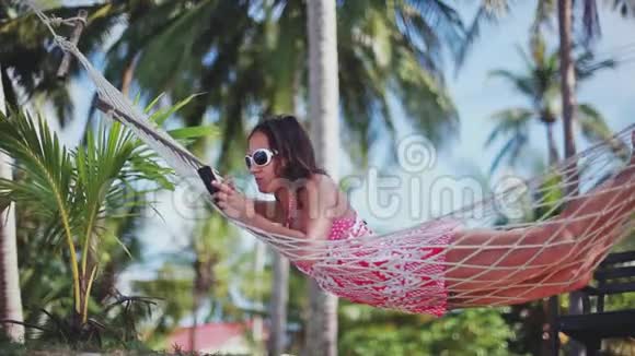 快乐海滩女人使用智能手机躺在吊床上放松热带度假1920x1080高清视频的预览图