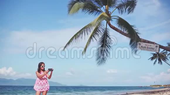 棕树下的沙滩上一位身穿红色连衣裙戴着墨镜带着手机拍照的美女视频的预览图
