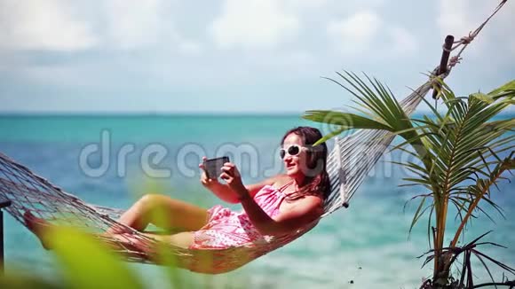 快乐的海滩女人戴着太阳镜带着手机自拍躺在吊床上在热带放松视频的预览图