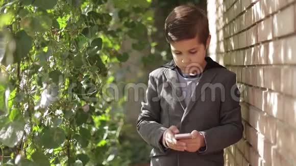 穿夹克的可爱的小男孩用他的手机常春藤墙的背景视频的预览图