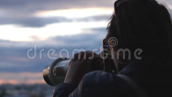 旅行者年轻的女游客在日落时用投币式望远镜观察城市并用手机拍照视频的预览图