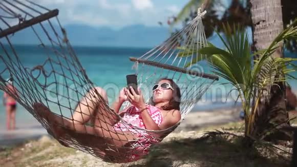 年轻的黑发女人戴着太阳镜在智能手机上发短信躺在吊床上放松热带度假视频的预览图