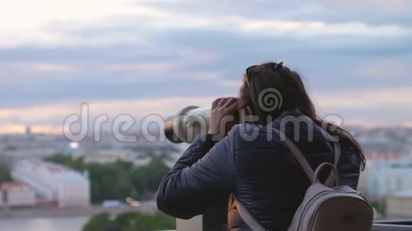 年轻的女游客带着背包在日落时分用投币式双筒望远镜缓慢地观察城市视频的预览图