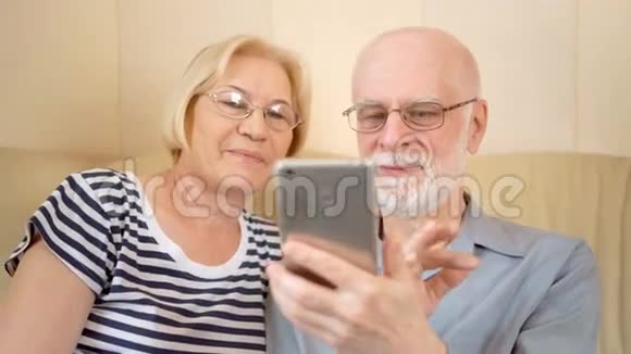 开朗好看的老两口坐在家里的沙发上通过Skype和智能手机聊天视频的预览图