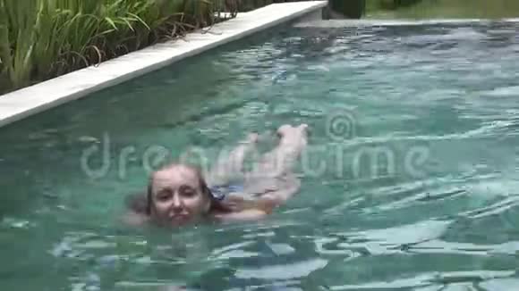 在热带度假胜地的棕榈树下比基尼有一头长发的年轻苗条女子在游泳池里游泳视频的预览图