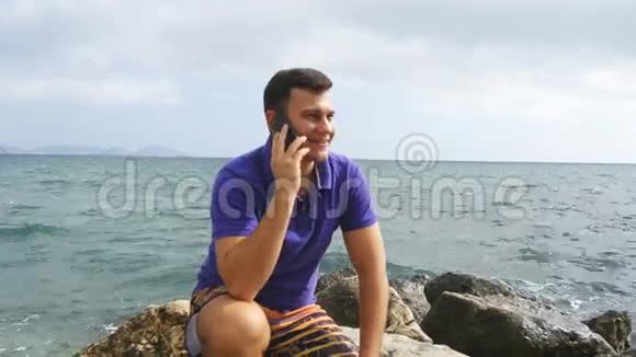 微笑的年轻人正在海边的海滩上用手机说话坐在海边的石头上的英俊快乐的家伙视频的预览图