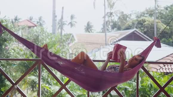 美丽的黑发女人在暑假期间在宁静的花园里放松在吊床上看书视频的预览图