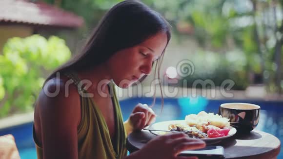 漂亮的女人坐在热带别墅度假村的露台上享受早餐使用手机异国情调视频的预览图