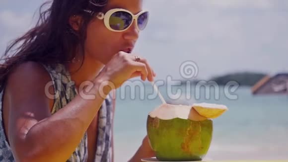 美丽的黑发女人戴着太阳镜在海滩上享受有趣的假期喝着新鲜的椰子水和稻草慢慢视频的预览图