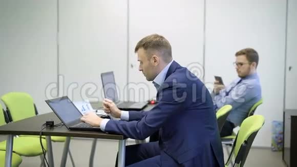 穿着带笔记本电脑的休闲服的年轻商人正坐在教室黑桌子后面的绿色椅子上一个视频的预览图