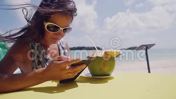 年轻的黑发美女戴着太阳镜在海滩上聊天喝着椰子做的鸡尾酒视频的预览图