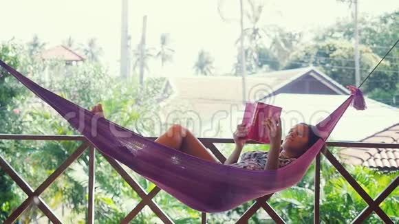快乐的年轻女性在暑假期间在一个宁静的花园里放松在一个摇摆的吊床上读一本书慢慢视频的预览图