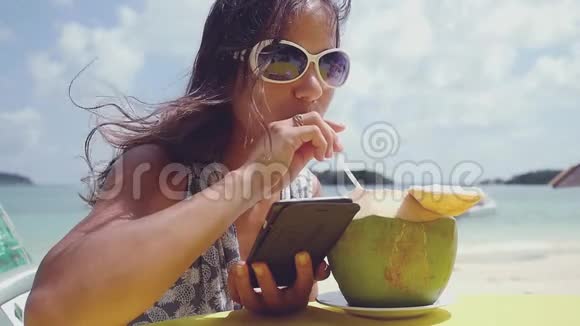 年轻漂亮的黑发女人戴着太阳镜在海滩上用椰子送信息喝鸡尾酒慢吞吞视频的预览图
