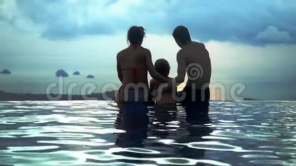 爸爸妈妈抱着儿子坐在水池边在山顶上看到无限的海景动作缓慢视频的预览图