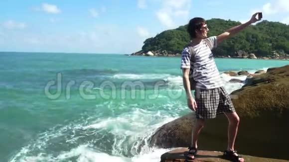 快乐的男人戴着太阳镜和背包在岩石海滩上用智能手机拍照苏梅岛3840x2160视频的预览图