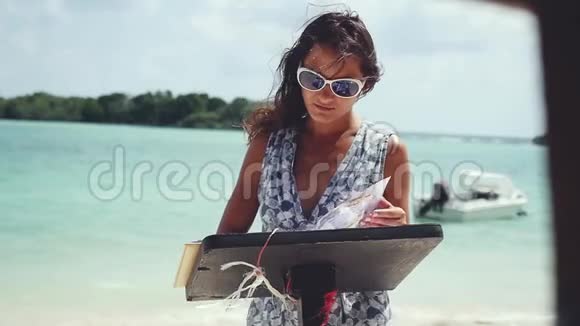 年轻的黑发女人戴着太阳镜在海滩餐厅看菜单她身后有船的大海景色视频的预览图