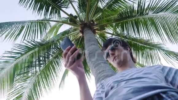 戴太阳镜的年轻人在棕榈树下户外使用移动智能手机3840x2160视频的预览图