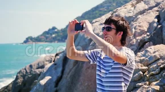 戴太阳镜的年轻人在岩石海滩用他的智能手机拍照慢动作苏梅岛泰国视频的预览图