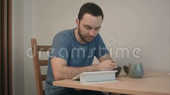 年轻人一边吃早餐一边在平板电脑上看新闻视频的预览图