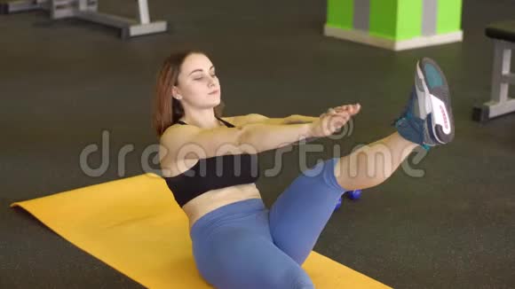 这名年轻女子躺在运动垫上在健身房里用手和腿做腹肌锻炼视频的预览图