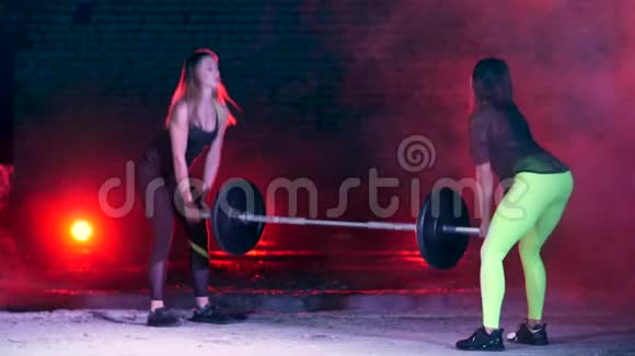 两个两人一组的运动女孩在晚上用杠铃在五颜六色的灯光下做各种力量练习视频的预览图