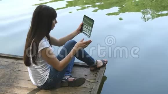 年轻快乐的女孩坐在湖边的码头上用平板电脑聊天视频的预览图