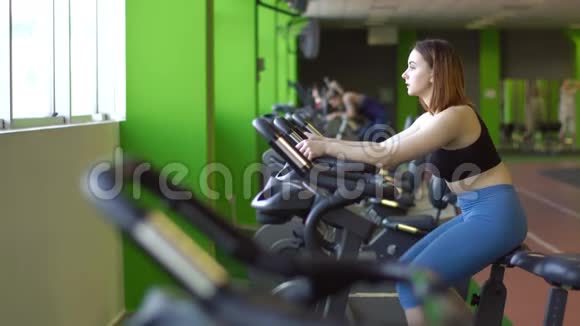 穿蓝色裤子和黑色上衣的妇女在绿色健身车上工作的横向肖像视频的预览图