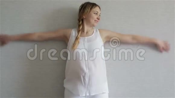 年轻的孕妇穿着一件白色上衣站在灰色的墙壁上幸福地挥动着他的手臂就像翅膀一样视频的预览图