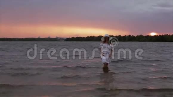 日落时分一个穿着连衣裙戴着太阳帽的中年妇女赤脚在水里散步视频的预览图