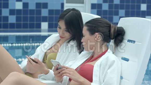 两个美女坐在泳池边的躺椅上看手机视频的预览图