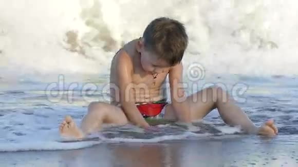 可爱的小宝宝坐在海滩上享受着和沙子玩耍的海浪慢动作视频的预览图