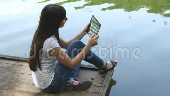年轻快乐的女孩坐在湖边的木制码头上在平板电脑上聊天视频的预览图