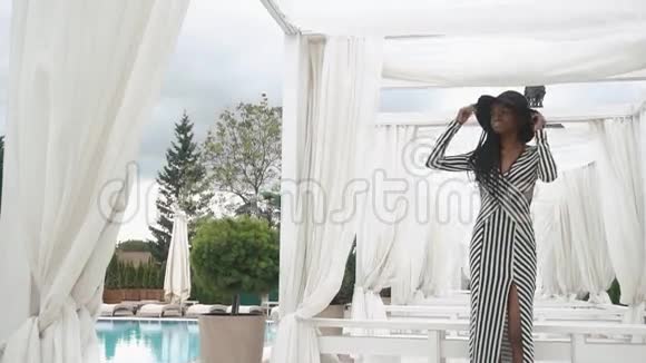 优雅的非洲裔美国女士穿着条纹鸡尾酒连衣裙和黑色帽子在豪华酒店背景下摆在游泳池附近视频的预览图